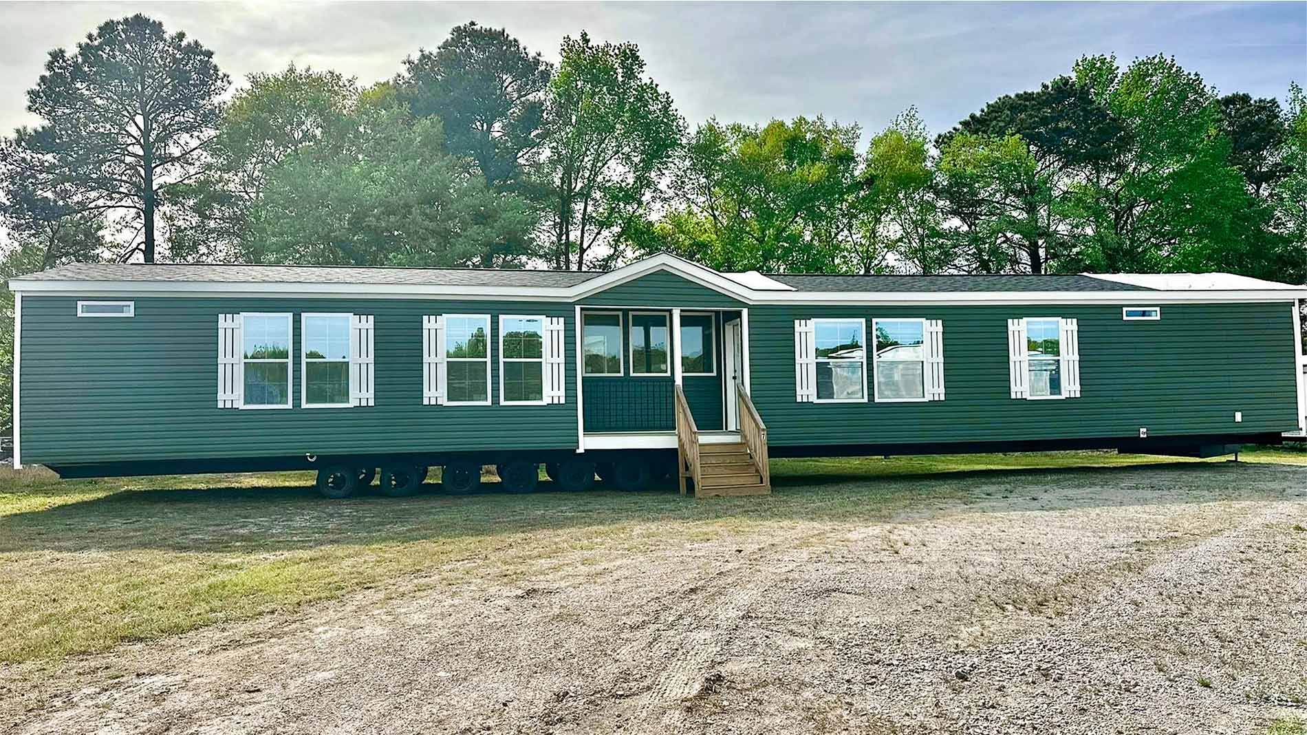 A spacious green manufactured home with white trim, multiple windows, and stairs sits on a gravel lot.