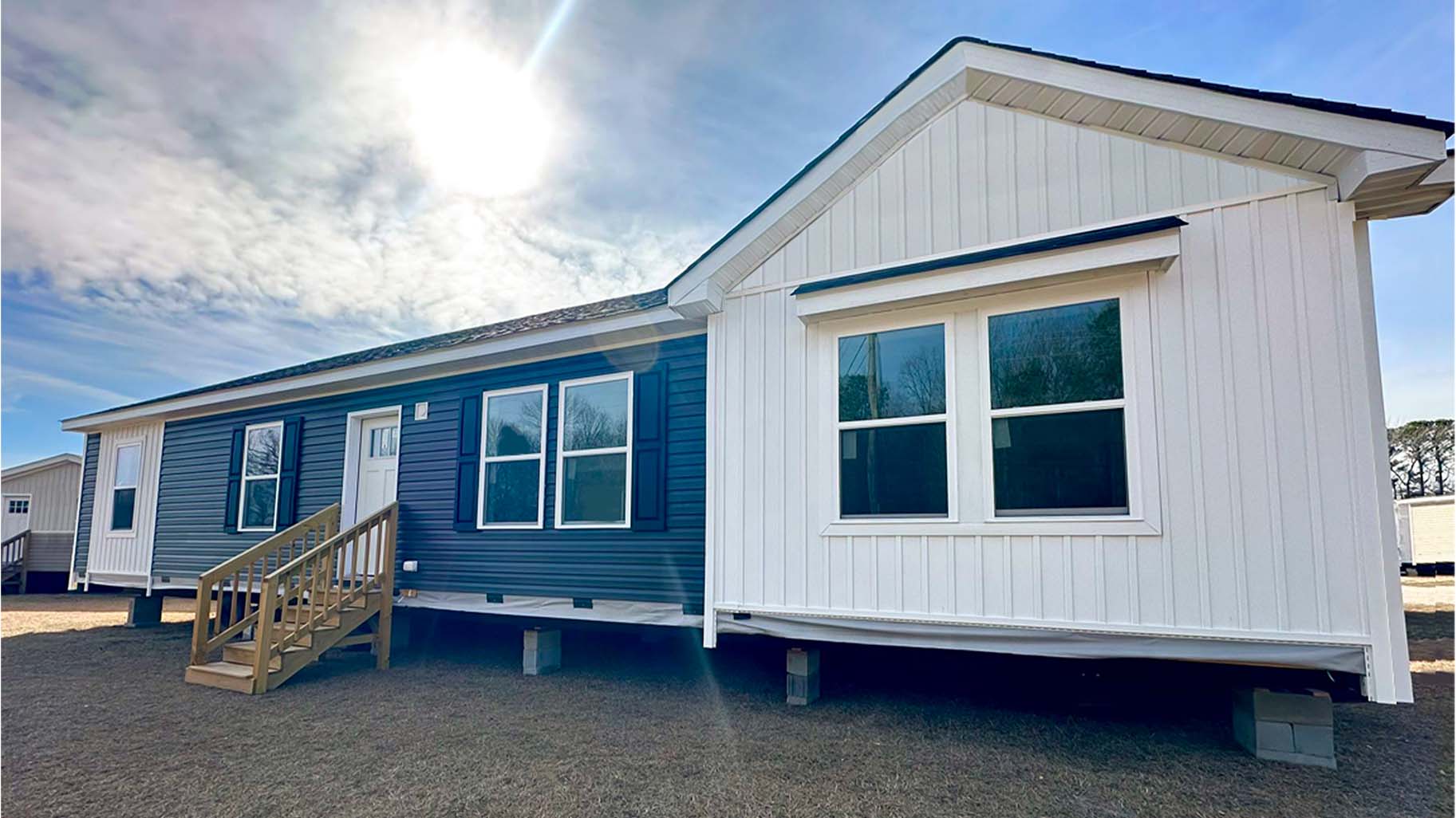 A modern modular home with white and blue siding, wooden steps, and large windows sits under a sunny sky.
